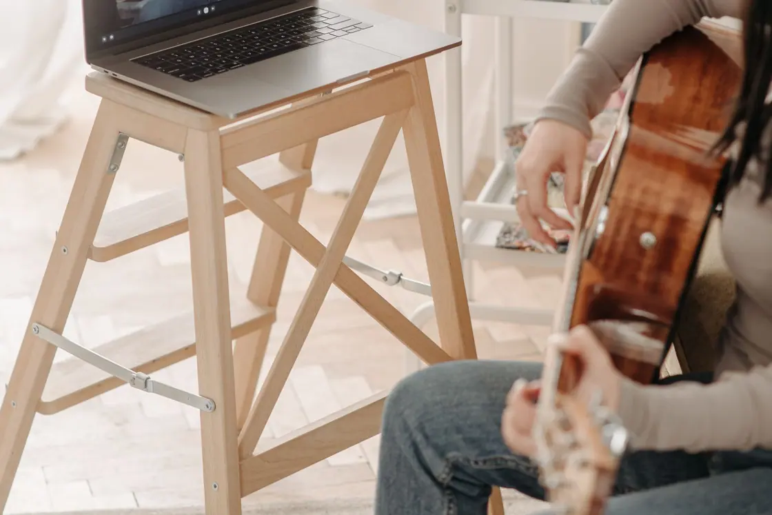An adult playing acoustic guitar beside a laptop at home
