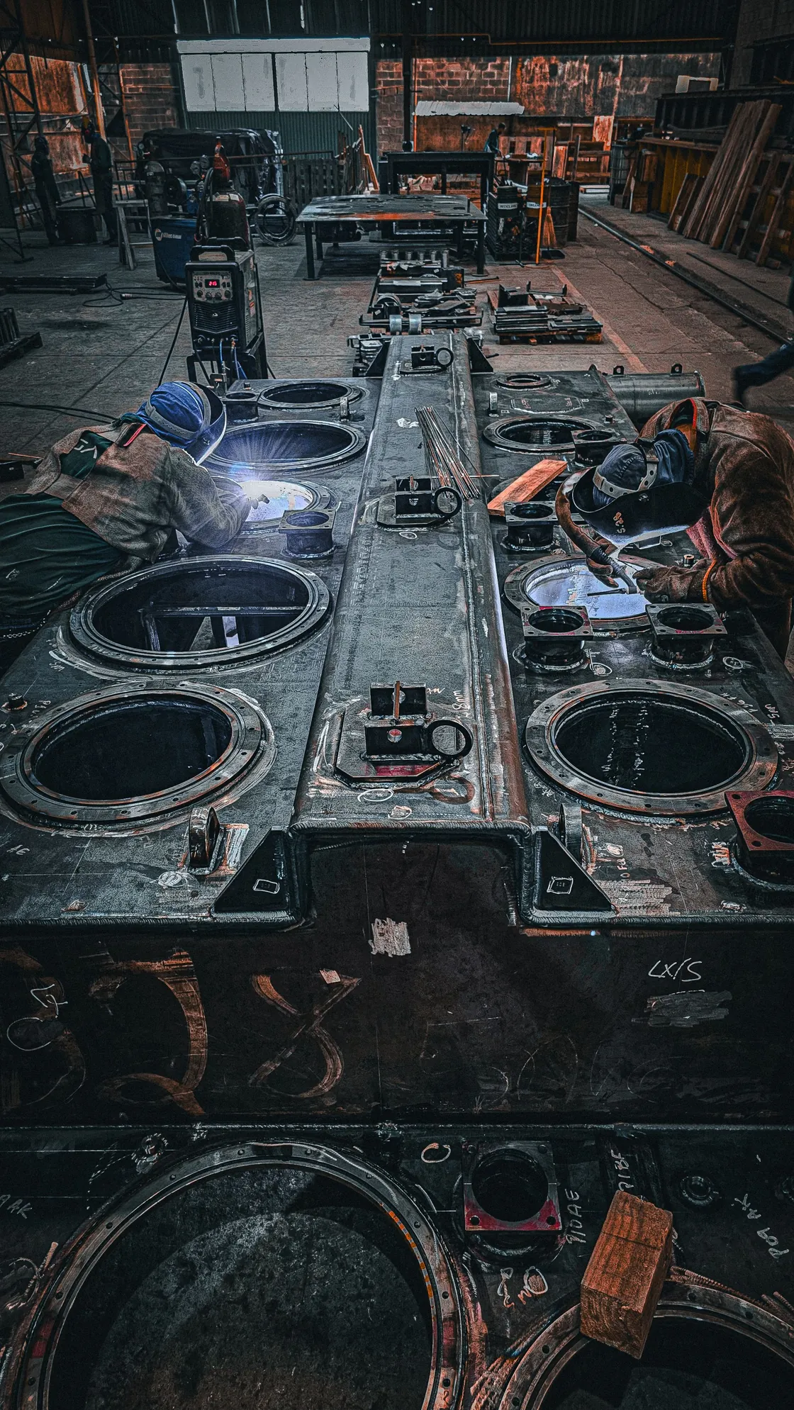 Two workers welding large metal components inside a factory