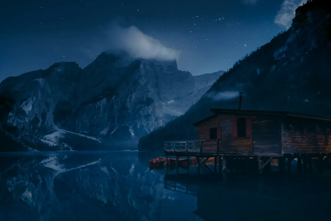 A lakeside boathouse and mountain reflected in deep blue water beneath a starry sky