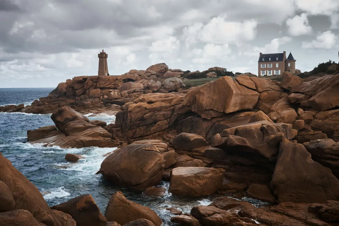 Pink granite rocks, a stone lighthouse and buildings on a grey coastal day