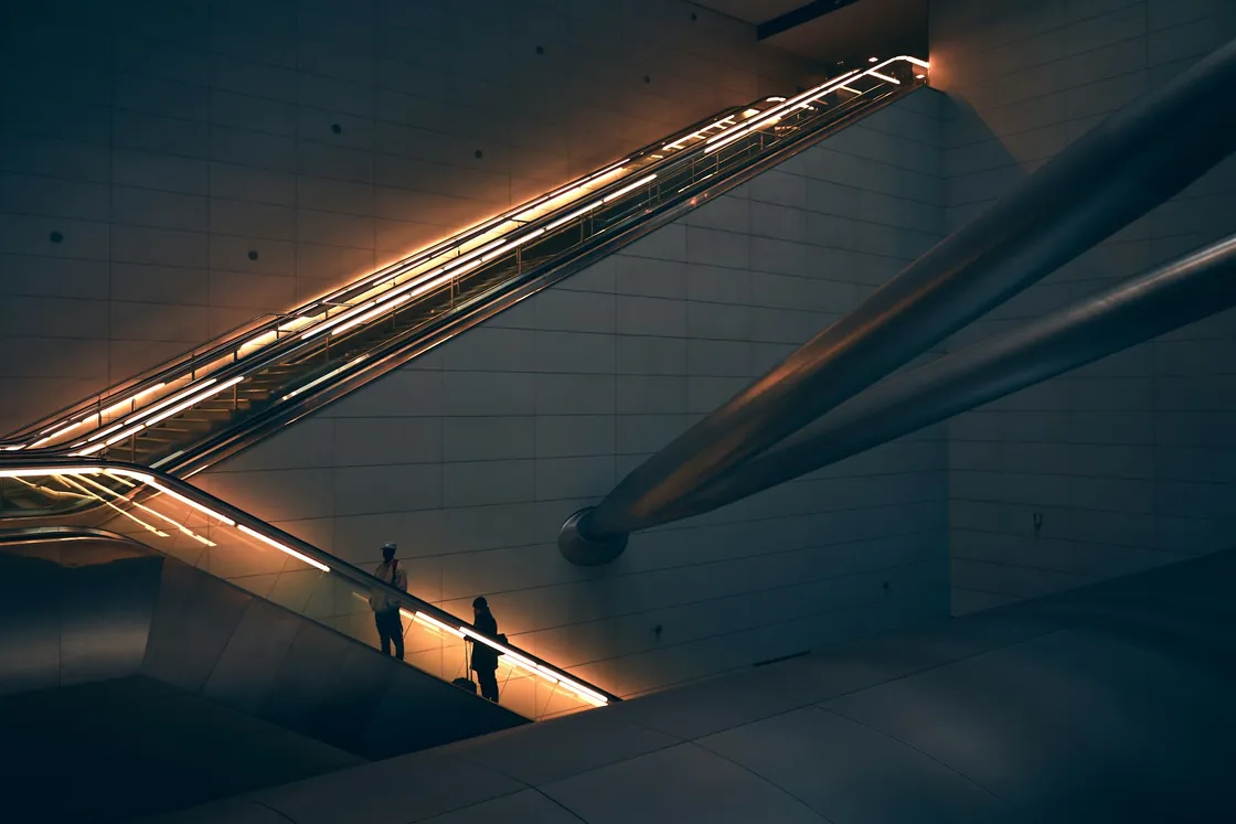 Two people on an illuminated escalator crossing the dark interior of a station