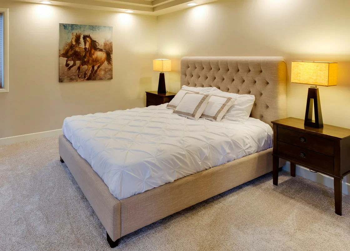 A double bed with a tufted beige headboard, warm bedside lamps and a horse painting