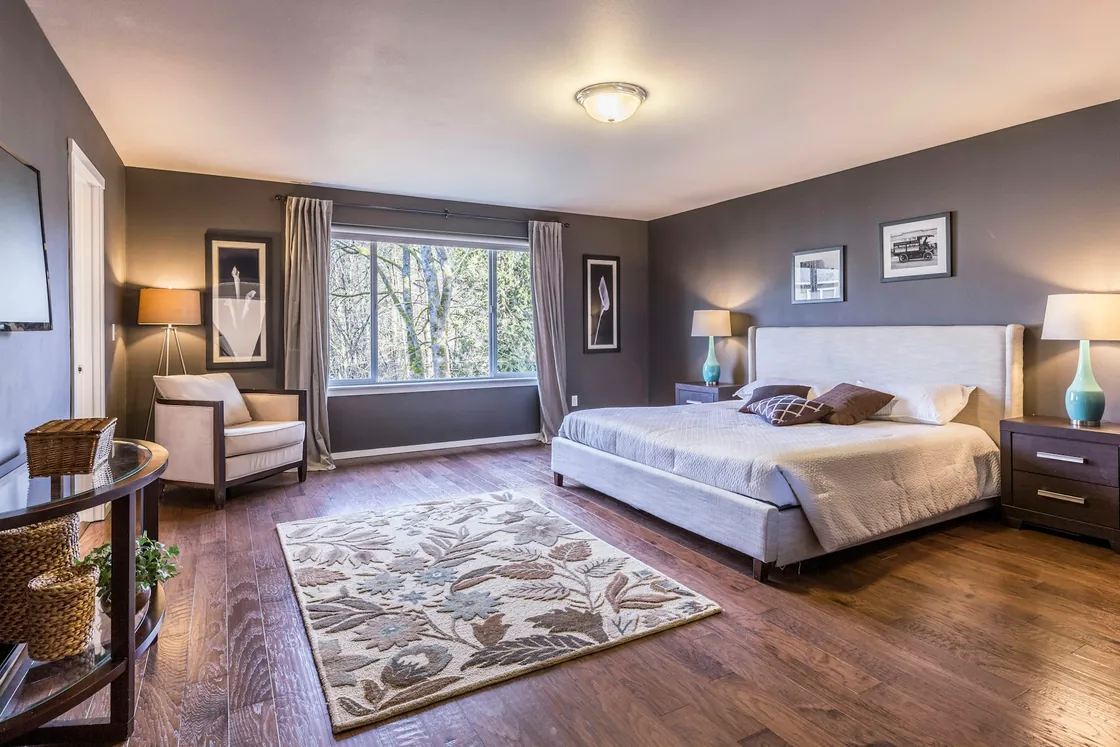 A spacious bedroom with grey walls, a large bed, an armchair and a window looking onto trees